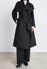 Dunkelgrauer Wollmantel mit Wickeldesign, der einen Bindegürtel in der Taille und weite Ärmel hat. Getragen über einem grauen Pullover und schwarzen Hosen.