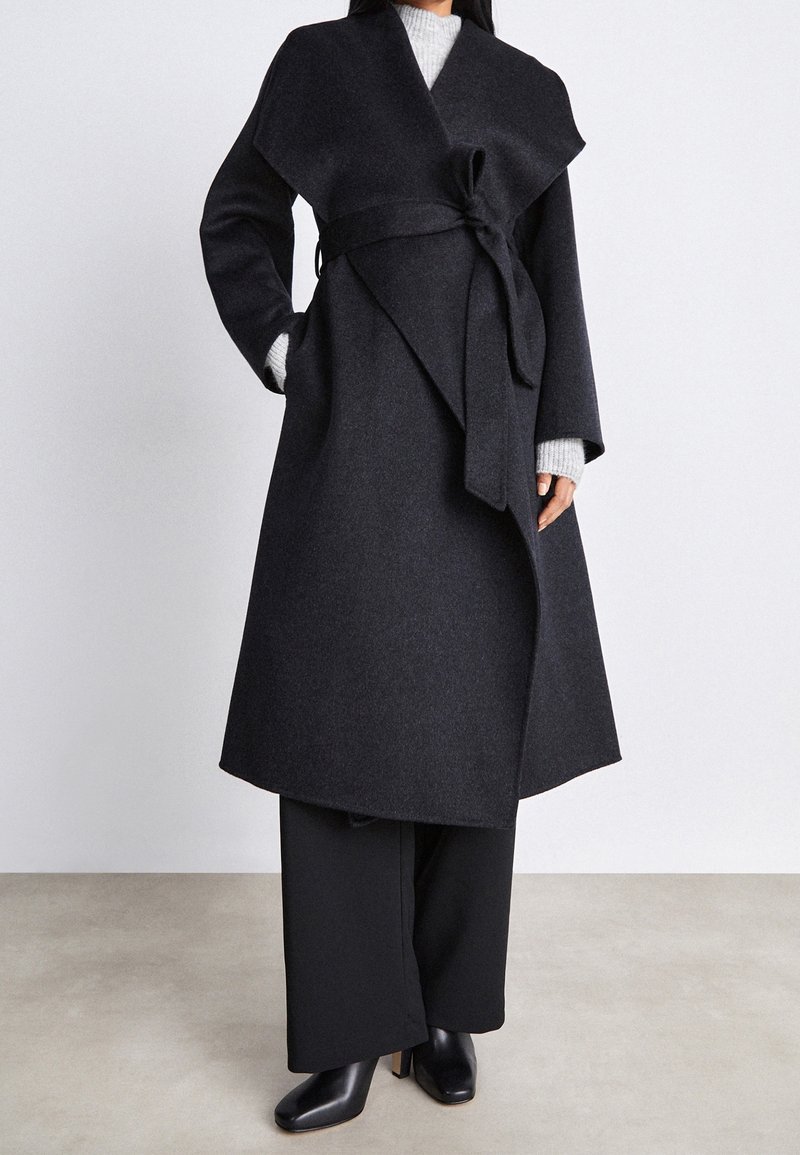 Dunkelgrauer Wollmantel mit Wickeldesign, der einen Bindegürtel in der Taille und weite Ärmel hat. Getragen über einem grauen Pullover und schwarzen Hosen.