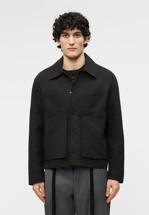 Νεαρός άνδρας με σγουρά σκούρα μαλλιά φοράει υφασμένο μαύρο σακάκι με κουμπιά και γκρι παντελόνι με ρίγες και μαύρους κάθετους ιμάντες.