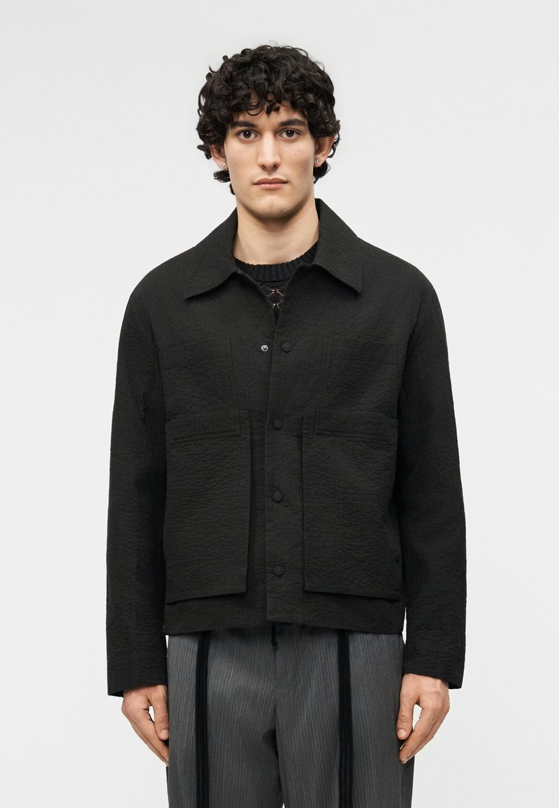 Fiatal férfi göndör, sötét hajjal, texturált fekete gombos dzsekit és szürke, függőleges fekete pántokkal díszített szürke csíkos nadrágot visel.