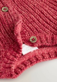 Gilet en tricot rouge avec un motif texturé, deux boutons ronds et une étiquette avec le nom de la marque partiellement visible au col.