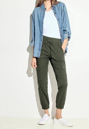 Mujer vestida con pantalones cargo de color verde oliva, zapatillas blancas, top blanco y camisa vaquera desabrochada con las mangas remangadas, de pie contra una pared blanca.