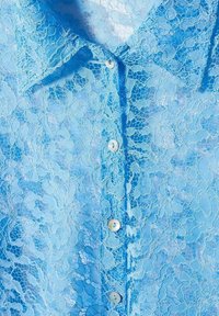 Chemise en dentelle bleu clair avec un col et une fermeture à boutons. Le motif en dentelle délicat crée un effet texturé et superposé.