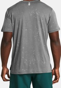 Šedé sportovní tričko s texturovaným povrchem, krátkými rukávy, bočními rozparky a třemi bílými akcentními pruhy na zadní části krku.