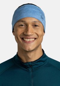 Diadema elástica de color azul claro con un acabado texturizado, que presenta un pequeño logo. Usada por una persona sonriente, que complementa una blusa de tono verde azulado oscuro.