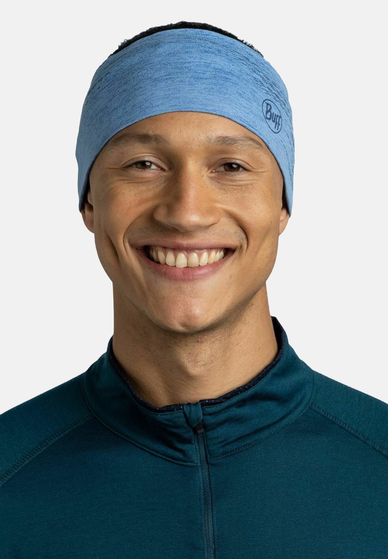 Diadema elástica de color azul claro con un acabado texturizado, que presenta un pequeño logo. Usada por una persona sonriente, que complementa una blusa de tono verde azulado oscuro.