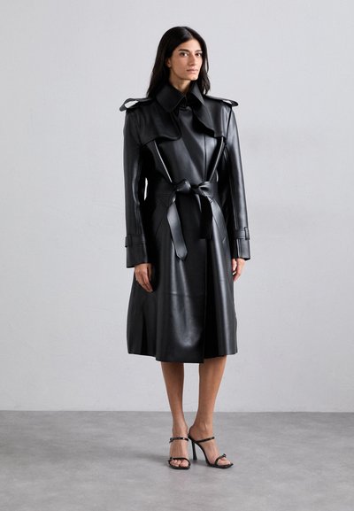 Sort læder trench coat med stor krave, bælte i taljen og skulderdetaljer. Glat tekstur og knælangt design. Hæl sandaler synlige.