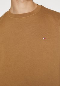T-shirt in cotone marrone con collo rotondo, caratterizzato da un piccolo logo ricamato in rosso, bianco e blu sul lato sinistro del petto.