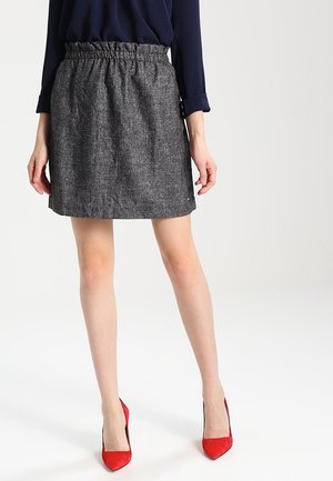 Vrouw die een grijze gestructureerde rok, een marineblauw shirt met lange mouwen en rode hoge hakken draagt, staand op een witte achtergrond.