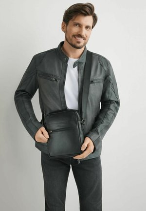 Mężczyzna uśmiechający się, ubrany w czarną skórzaną kurtkę i ciemne jeansy, trzymający czarną torbę na ramię na jednolitym jasnym tle.