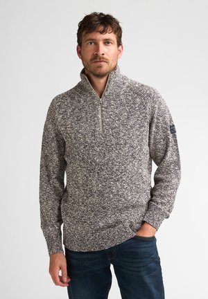 Mann mit kurzen braunen Haaren und Bart, trägt einen grau-weißen Strickpullover mit Viertel-Reißverschluss und dunkelblauen Jeans, steht vor einem schlichten Hintergrund.