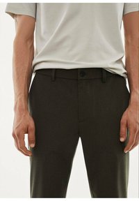 Dark green trousers with a smooth texture, featuring a button closure and belt loops. Paired with a light grey t-shirt, showing hands on hips.
