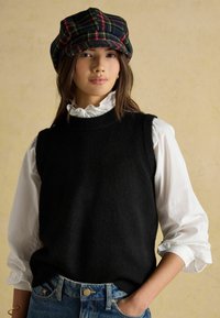 Czarny bezrękawnik z teksturowanym splotem, noszony na białej bluzce z falbankami. W połączeniu z kratką płaską czapką. Niebieskie dżinsy jeansowe widoczne w talii.