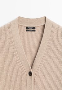 Beige vlněný kardigan s výstřihem do V, ribovanou texturou a jedním hnědým knoflíkem na zapínání. Na štítku je nápis "Massimo Dutti, 100% vlna."