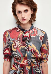 Blusa stampata in colori vivaci con motivi a spirale e floreali. Realizzata in tessuto leggero, con colletto annodato e maniche corte a sbuffo.