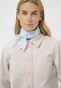 Bufanda azul claro atada al cuello, con una sutil textura de patrón. El modelo lleva una camisa beige de botones con un bolsillo en el pecho.