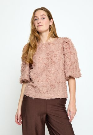 Žena s dlouhými vlasy, oblečená v texturovaném prachově růžovém tričku s krátkými rukávy a volných hnědých kalhotách, stojící před čistým bílým pozadím.