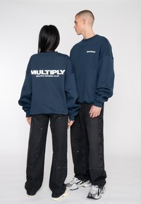 Felpe blu navy con testo bianco "MULTIPLY APPAREL CLUB", abbinate a pantaloni neri a gamba larga. Vestibilità casual e tessuto testurizzato.