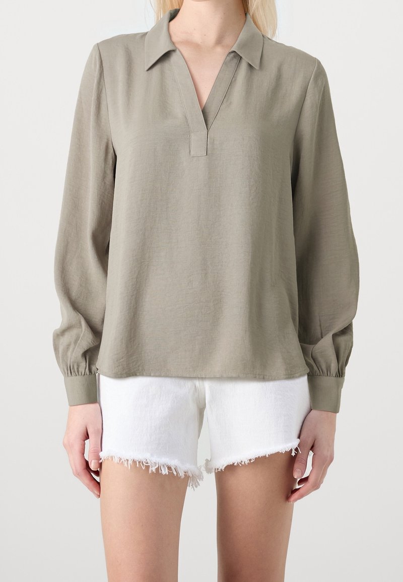 Kvinna iklädd en lös olivgrön långärmad blus med V-halsringning och vita denim-shorts med fransar.