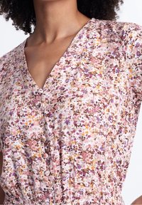 Abito con stampa floreale caratterizzato da scollatura a V, chiusura con bottoni e vita elastica. I materiali mostrano tonalità di fiori rosa, viola e giallo su uno sfondo chiaro.