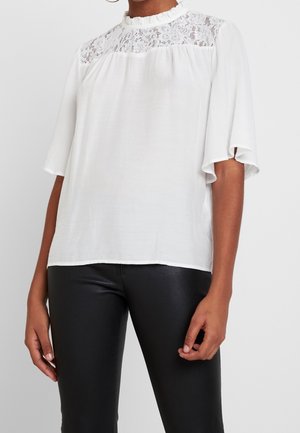 Frau trägt eine weiße Bluse mit Spitzenpasse und Rüschenkragen, kombiniert mit schwarzen, eng anliegenden Hosen, stehend vor schlichtem Hintergrund.