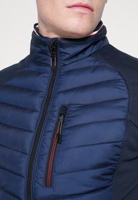 Mann trägt eine marineblaue gesteppte Jacke mit hohem Kragen, schwarzem Reißverschluss und einer mit roten und weißen Akzenten versehenen, mit Reißverschluss verschließbaren Brusttasche.