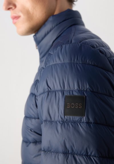 Homme portant une veste matelassée bleu marine avec un col et un patch logo BOSS noir sur la manche supérieure, tourné vers la gauche.