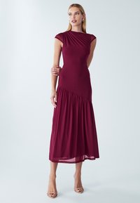 Nainen seisoo kädet ristissä yllään sanssiton viininpunainen midi-mekko, jossa on pleat-tyyppiset yksityiskohdat, ja hänellä on nude-väriset avonaiset kengät tasaisella taustalla.