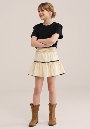 Junges Mädchen lächelt mit verschränkten Armen, trägt ein schwarzes T-Shirt, einen cremefarbenen Rock mit schwarzem Rand und braune Cowboy-Stiefel vor einem schlichten Hintergrund.