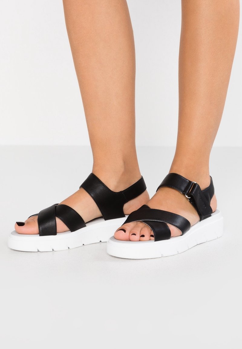Pieds portant des sandales en cuir noir avec des semelles blanches épaisses et des brides à la cheville sur un fond clair.