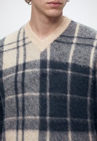 Jeune personne portant un pull en maille beige et gris foncé à carreaux avec un col en V, visible des épaules au menton.