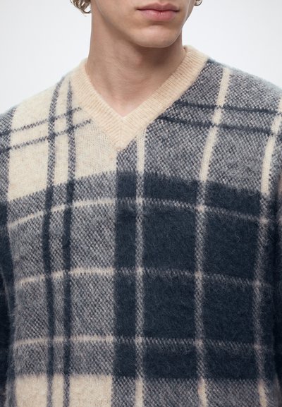 Jeune personne portant un pull en maille beige et gris foncé à carreaux avec un col en V, visible des épaules au menton.