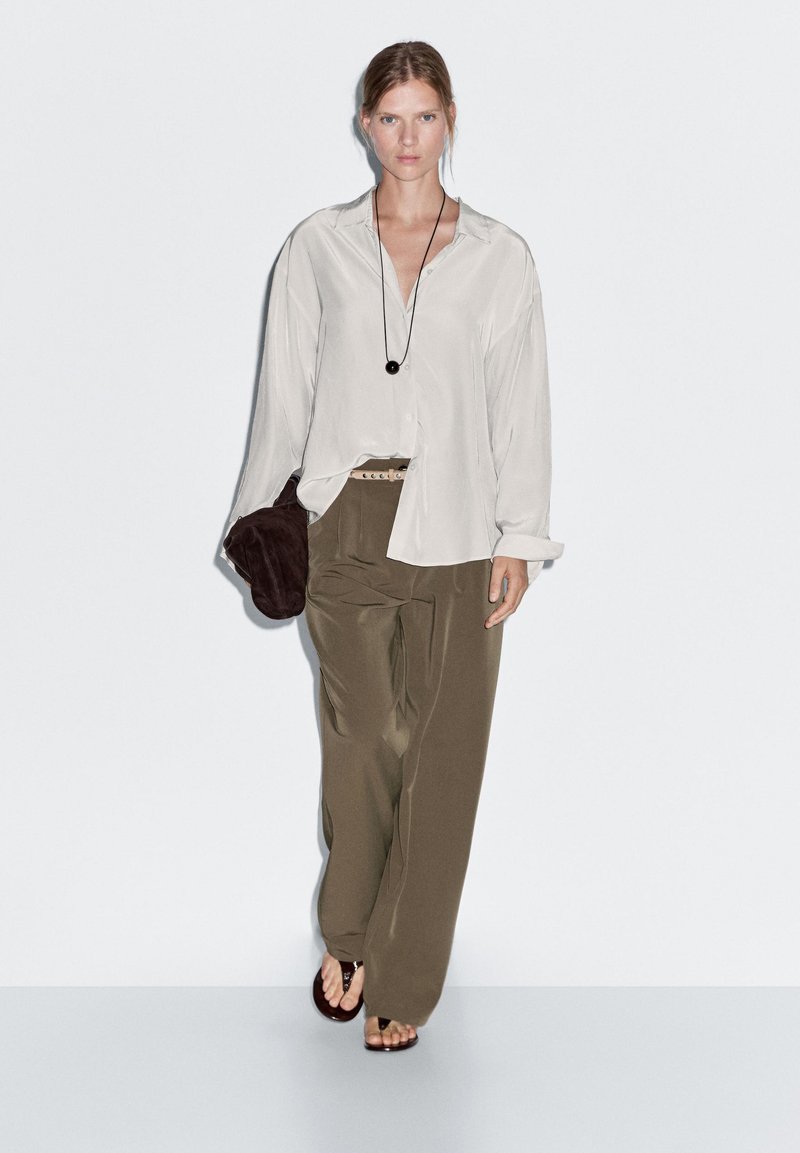 Helles beige Hemd mit Knöpfen und langen Ärmeln, olivgrüne, weite Hose, und eine braune Clutch; schwarze Halskette mit einem runden Anhänger.