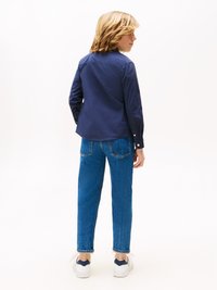 Marinblå skjorte med lange ermer, blå denimjeans med falmet tekstur, og hvite sneakers med marineblå detaljer. Sett fra baksiden.