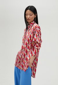 Patterned shirt with red, blue, and cream geometric shapes; long sleeves, button-down collar, and relaxed fit, paired with blue trousers.