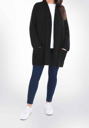 Femme portant un long cardigan noir avec poches sur une chemise blanche, un jean skinny bleu foncé et des baskets blanches, debout sur un fond blanc.