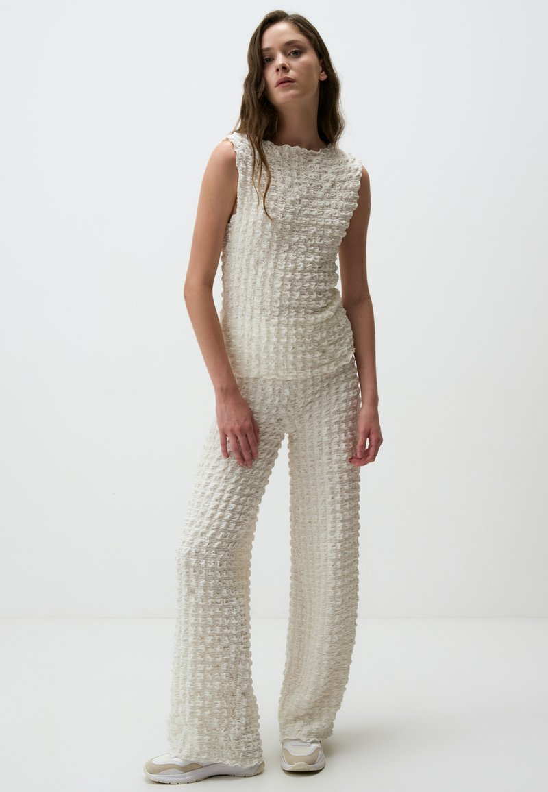 Weißes, strukturiertes Strickoutfit mit einem ärmellosen Top und weit geschnittenen Hosen. Der Stoff hat ein erhabenes Muster, das durch hellfarbige Schuhe ergänzt wird.