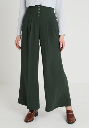 Spodnie z wysokim stanem i szerokimi nogawkami wykonane z ciemnozielonej tkaniny, z przodu z zakładkami i pięcioma dużymi guzikami. Całość uzupełniają brązowe mokasyny.