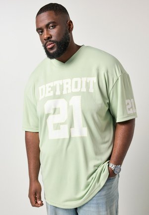 Hombre con barba que lleva una camiseta deportiva Detroit 21 verde claro y vaqueros azul claro, mirando a la cámara con expresión neutra.