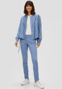 Svijetloplavi bomber jakna sa zipom sprijeda i naboranim rukavima, uparen sa svijetloplavim slim-fit trapericama i bijelim tenisicama. Gladak materijal.