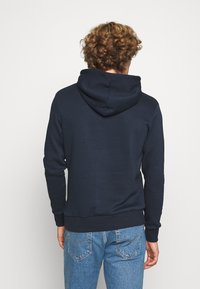 Marinblå huvtröja med mjuk textur, utrustad med känguruficka och ribbade manschetter. Bakifrån visas en avslappnad passform.