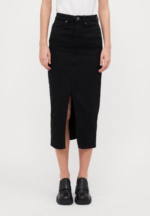 Kobieta w czarnej, midi spódnicy z wysokim stanem i przednim rozcięciem oraz czarnych loafersach, zestawiona z białą bluzką.