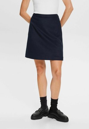 Vrouw draagt een zwarte rok tot op de knie, wit topje, zwarte sokken en zwarte loafers, staat met handen achter haar rug.