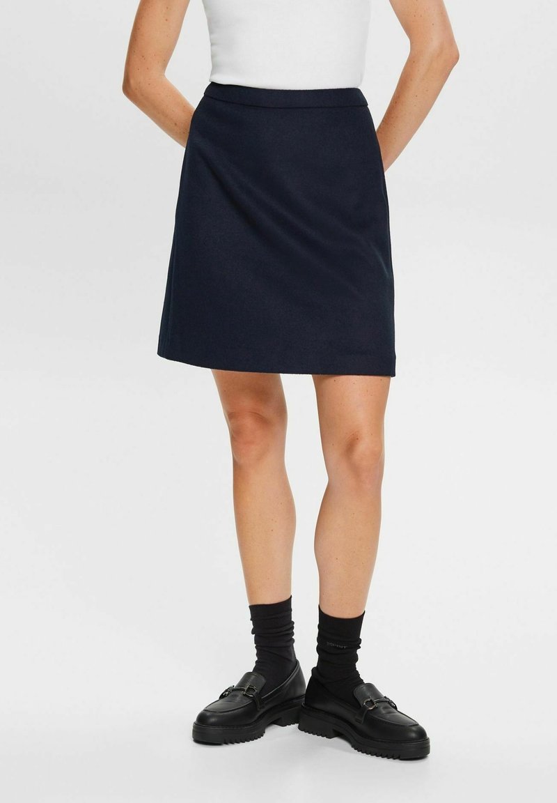Femme portant une jupe noire mi-longue, un haut blanc, des chaussettes noires et des mocassins noirs, debout les mains derrière le dos.