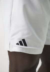 Shorts de sport blancs en tissu texturé, arborant un logo Adidas noir avec trois bandes sur le coin inférieur gauche.