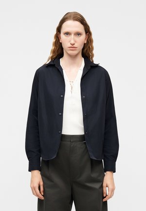 Žena s dlhými vlnitými vlasmi, oblečená v čiernej bunde na gombíky cez biely horúci top so šnurovaním a tmavé nohavice s vysokým pásom, stojaca pred jednoliatym pozadím.