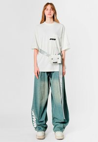 Camiseta blanca de gran tamaño con bolsillo delantero y letras negras, combinada con vaqueros sueltos de mezclilla azul desgastada y una riñonera blanca. Zapatos de plataforma blancos.