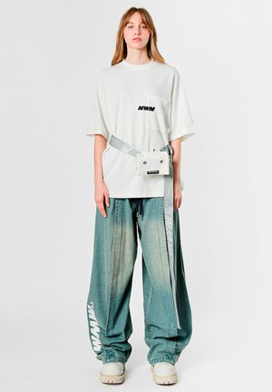 Camiseta blanca de gran tamaño con bolsillo delantero y letras negras, combinada con vaqueros sueltos de mezclilla azul desgastada y una riñonera blanca. Zapatos de plataforma blancos.