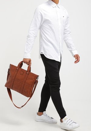 Sac en cuir marron avec deux poignées supérieures et une bandoulière, avec des accents cousus et une finition lisse. Porté par une personne en tenue décontractée.