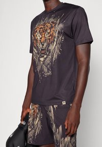 Man med mörk atletisk outfit med en rytande tiger graphic på tröjan och shortsen, som håller svarta boxhandskar i ena handen.